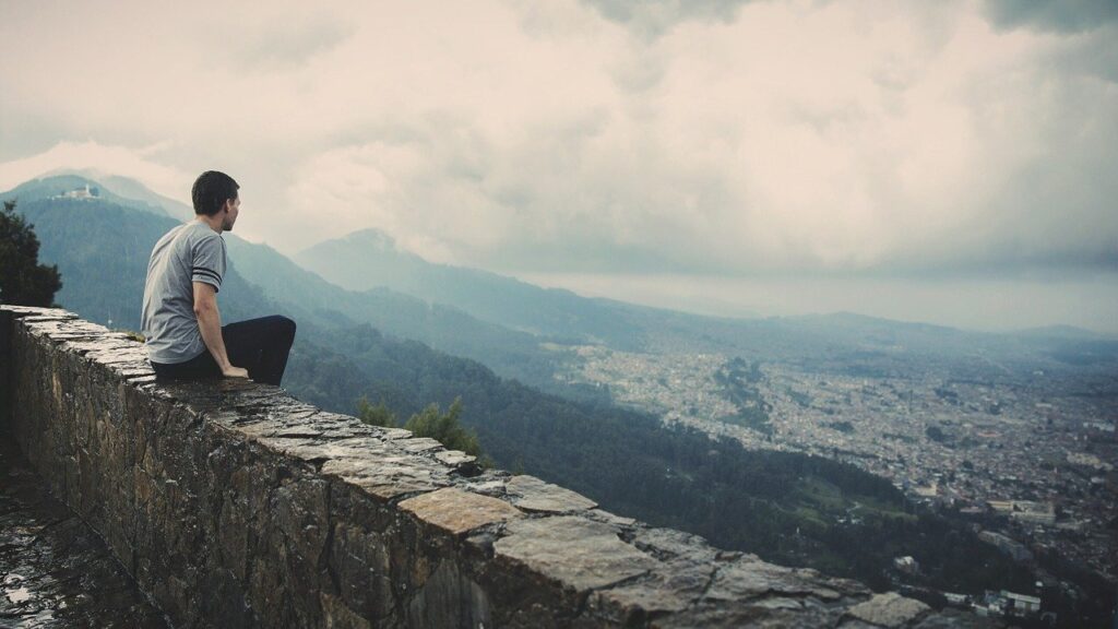 Un homme, assis sur un muret de pierre, contemple les montagnes et une ville au loin.